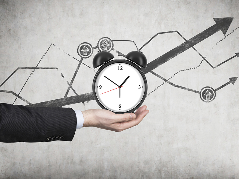 A businessman's hand holds an alarm clock. There is a growing line charts behind the alarm clock. A concept of time management or billing services in legal or consulting company.